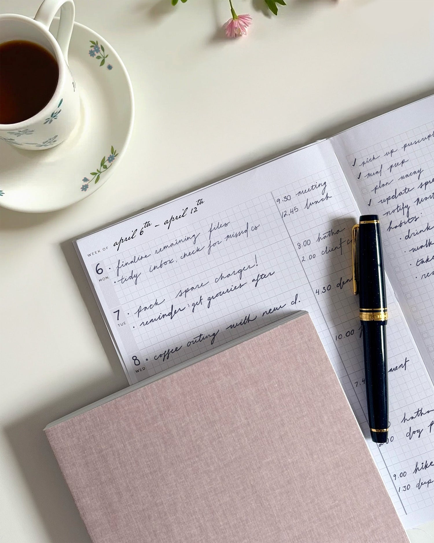 A top down view of a Hemlock and Oak cloth flex notebook in Aster, with a second open planner showing the handwritten plans. A closed fountain pen rests on the page and a cup of tea is off to the side.