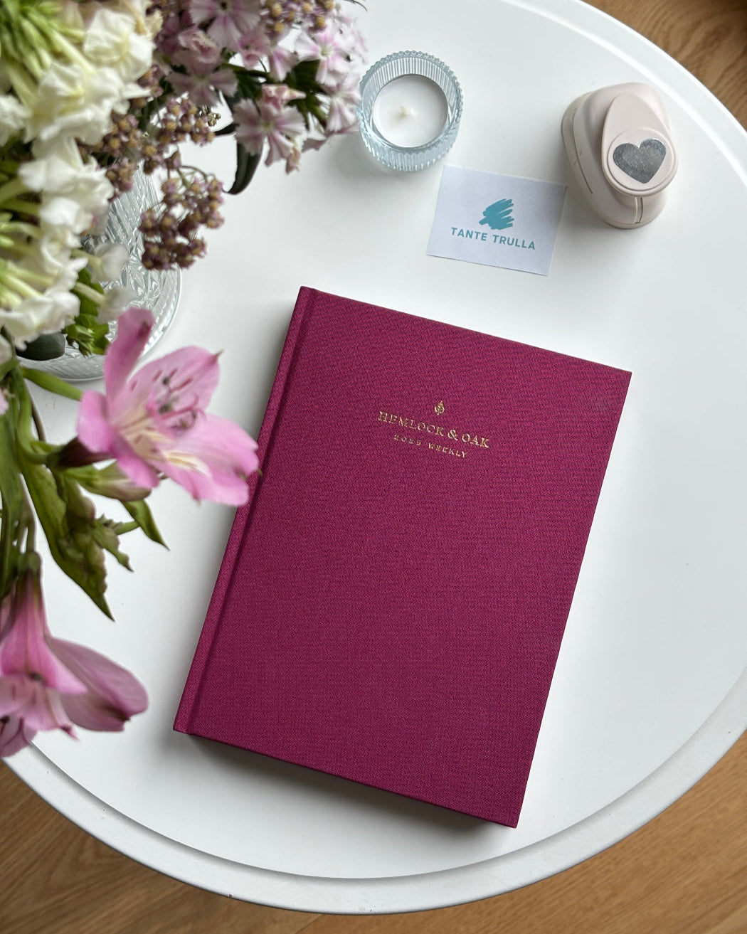 Purple notebook with a brand logo on a white table surrounded by flowers and a candle.