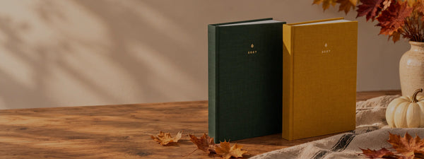Forest green and golden yellow 2027 planners on a wooden table with autumn leaves, linen and an ivory pumpkin