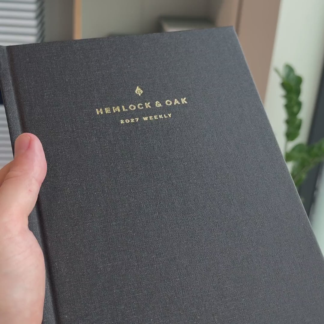 2027 Sunday Start Weekly Planner (Hardcover, Vertical, Heritage 120gsm, A5+) in Charbon, held in hand #color_charbon